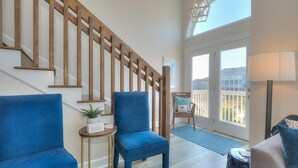 A staircase with wooden railings, a blue chair, a small table with a plant, and a view of the outside through a glass door. - Discover Blue Dolphin in Oak Island: a spacious 5-bedroom oceanview home for 12! (Oak Island)