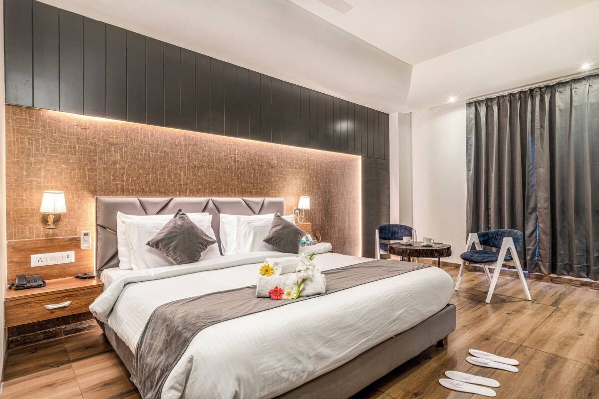 Habitación de hotel con una cama grande, mesitas de noche con lámparas, una pequeña mesa redonda, una silla azul y vistas al exterior a través de cortinas.