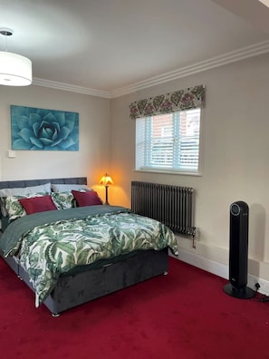 Une chambre à coucher avec un lit, un radiateur, une fenêtre avec des stores, une lampe fixée au mur et un grand tableau représentant des motifs floraux.