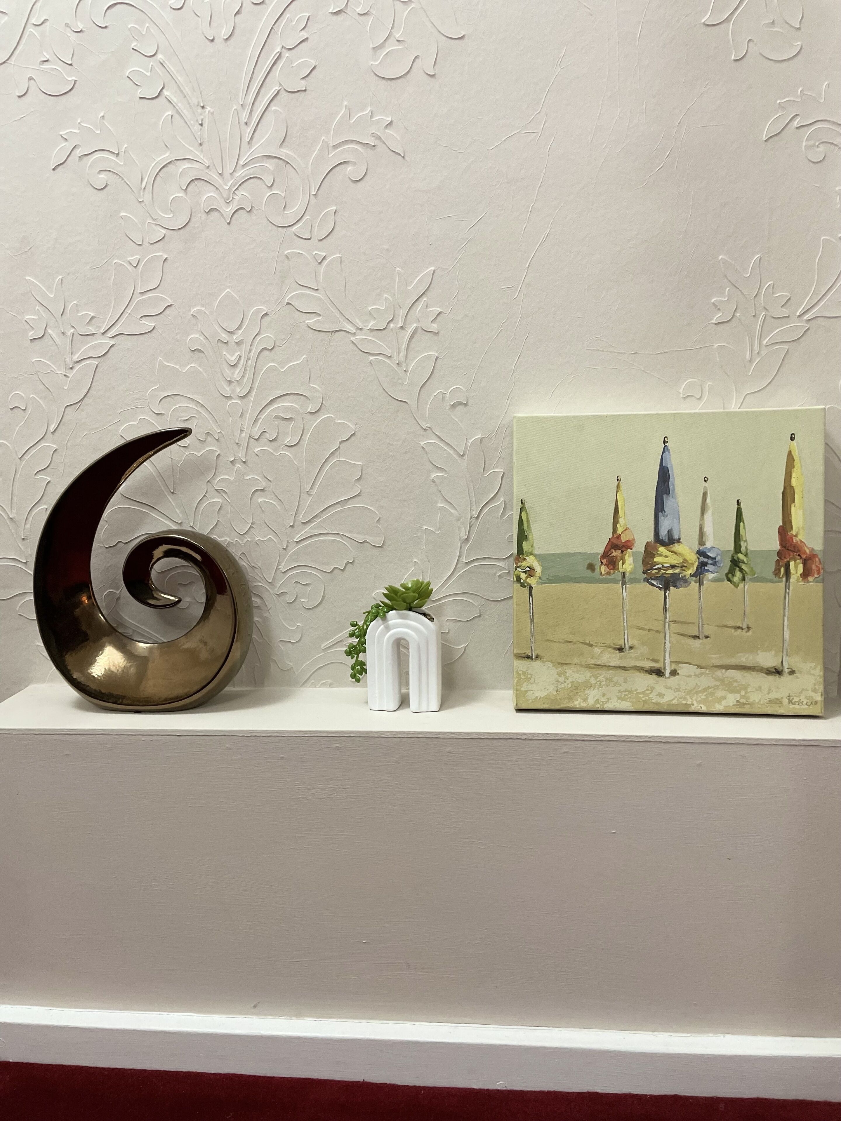 Una pared decorativa con motivos florales, una escultura en forma de luna creciente metálica, una maceta blanca pequeña con una suculenta y un cuadro de sombrillas de playa.