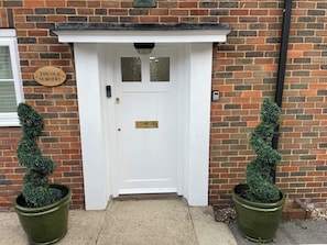 Une porte d’entrée blanche avec une plaque en laiton, une petite fenêtre au-dessus et une enseigne sur le mur, à gauche.