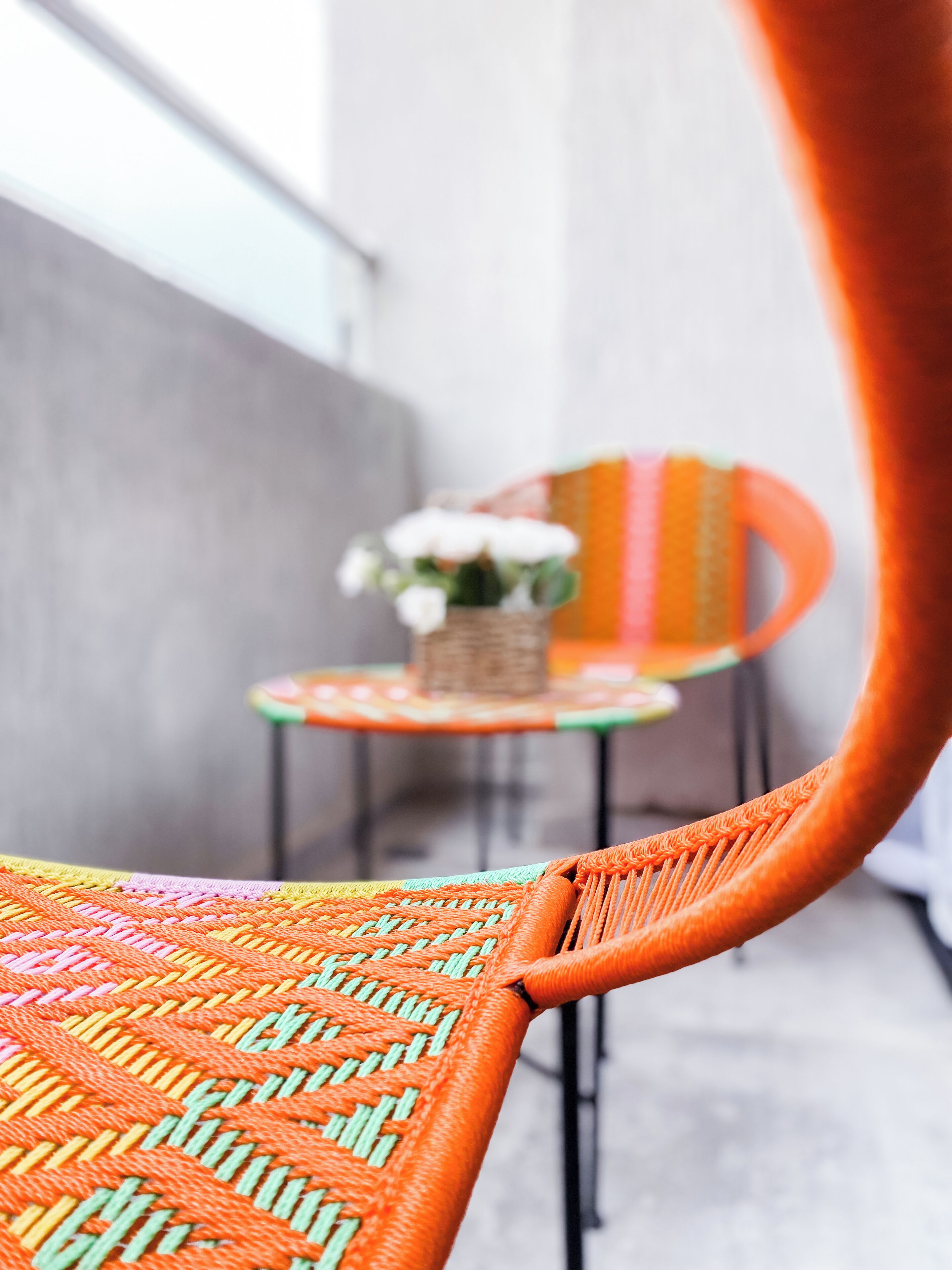 Una sedia in vimini con seduta e schienale intrecciati, un tavolino rotondo con un cesto in vimini e fiori bianchi, e una sedia colorata a righe sullo sfondo.