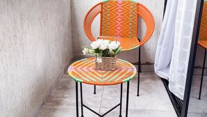 Un balcón con dos sillas naranjas, una pequeña mesa con una cesta tejida y flores, y vistas de edificios al fondo.