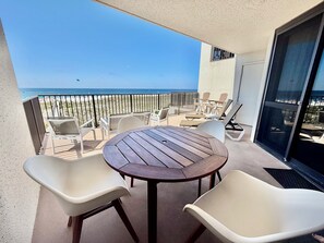 A balcony with a wooden table and white chairs, overlooking a beach and ocean.