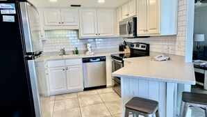 Una cocina moderna con armarios blancos, un refrigerador de acero inoxidable, un microondas, un horno y un lavavajillas. Hay dos taburetes en la isla.