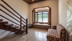 A room with a wooden staircase, a built-in bench with a patterned cushion, a sofa, and a view of a lake through a window. - Meghavan Resort By DLS Hotels (Dharamshala)