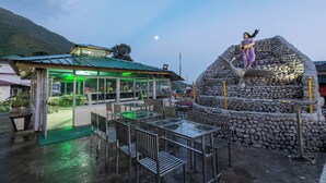 An outdoor seating area with tables and chairs, a stone sculpture, and a building with a green roof. - Meghavan Resort By DLS Hotels (Dharamshala)