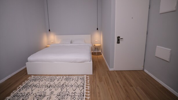 A bedroom with a white bed, a patterned rug, and a door with a light switch. - Stylish apartment in central TLV just a short walk from the beach (Tel Aviv)