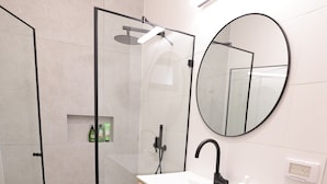 A modern bathroom with a glass-enclosed shower, a wooden vanity with a white sink, and a round mirror. - Stylish apartment in central TLV just a short walk from the beach (Tel Aviv)