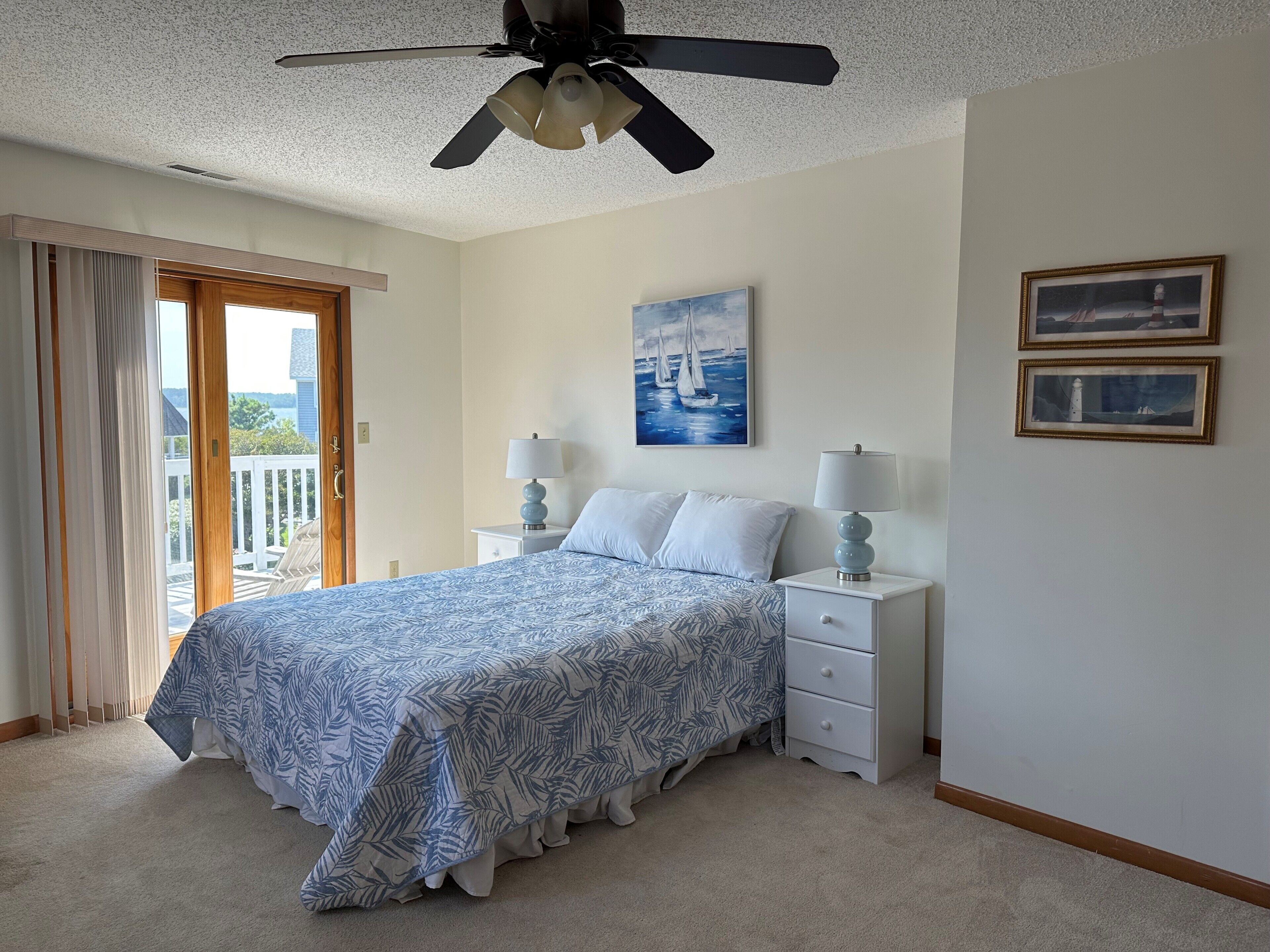 Une chambre à coucher avec un lit, deux tables de chevet, un ventilateur de plafond, un tableau et une vue sur l’extérieur, visible à travers une porte.