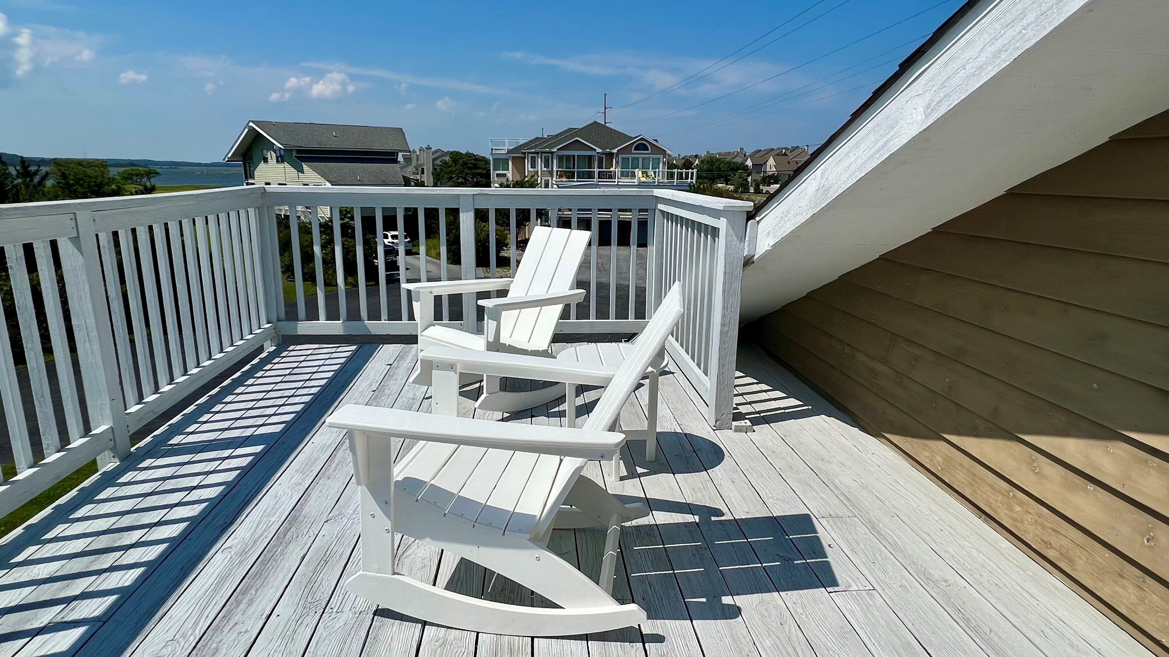 A wooden deck with white Adirondack chairs, a white railing, and a view of houses in the background.