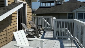 A wooden deck with white chairs and a small table, a covered area with a railing, and a building with a gabled roof in the background. - 5 Bedroom Dog Friendly House with community pool and beach views! (Fenwick Island)