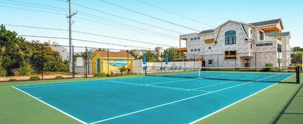 Uma quadra de tênis ao ar livre, com rede, cercada por uma grade e adjacente a um edifício com uma placa.