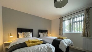 A hotel room with two beds, a window with curtains, a nightstand with a lamp, and a radiator.