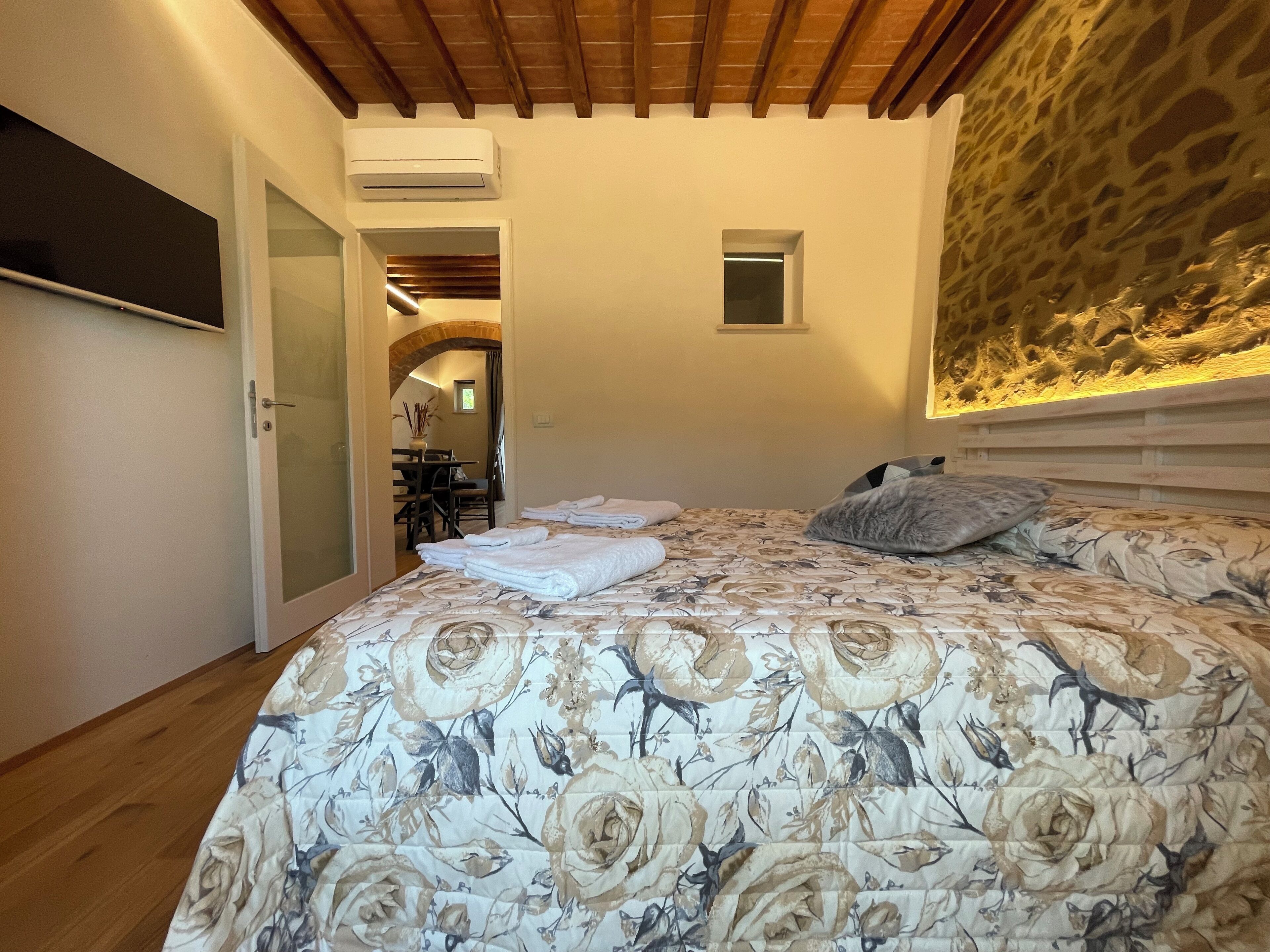 Een slaapkamer met een bloemenbedovertrek, een accentmuur van steen en een plafond met houten balken.