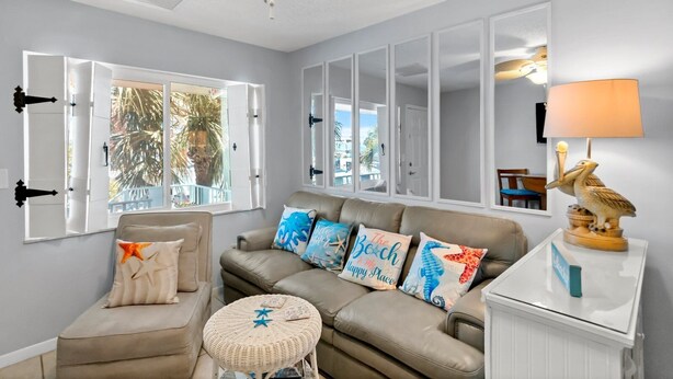 A living room with a beige sofa, a round wicker ottoman, and a coffee table. There are decorative pillows on the sofa and a pelican lamp on a side table. - Steps to Beach Access | Updated Condo | Pool | Sleeps 4 (Madeira Beach)