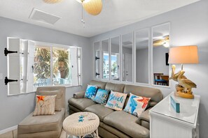 Ein Wohnzimmer mit einem beigen Sofa, einem runden Rattan-Pouff und einem Couchtisch. Auf dem Sofa liegen dekorative Kissen und auf einem Beistelltisch steht eine Lampe in der Form eines Pelikans.