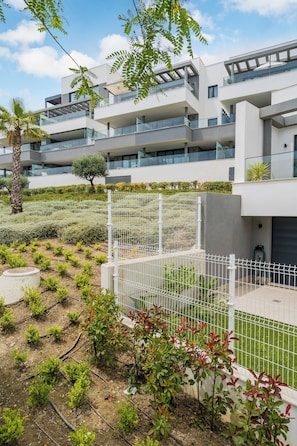Un moderno edificio residenziale a più piani con balconi, un'area giardino con piccole piante e una recinzione bianca.