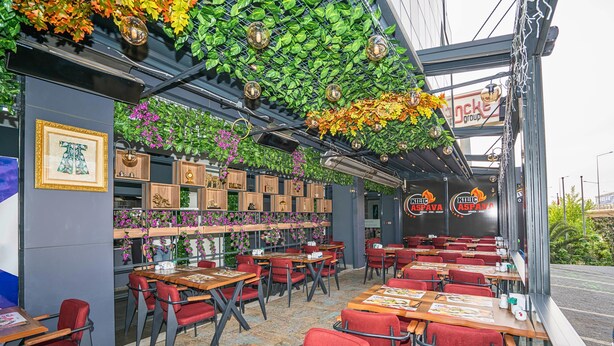 Restaurante interno com cadeiras vermelhas, mesas de madeira e um teto decorado com plantas e luminárias pendentes.
