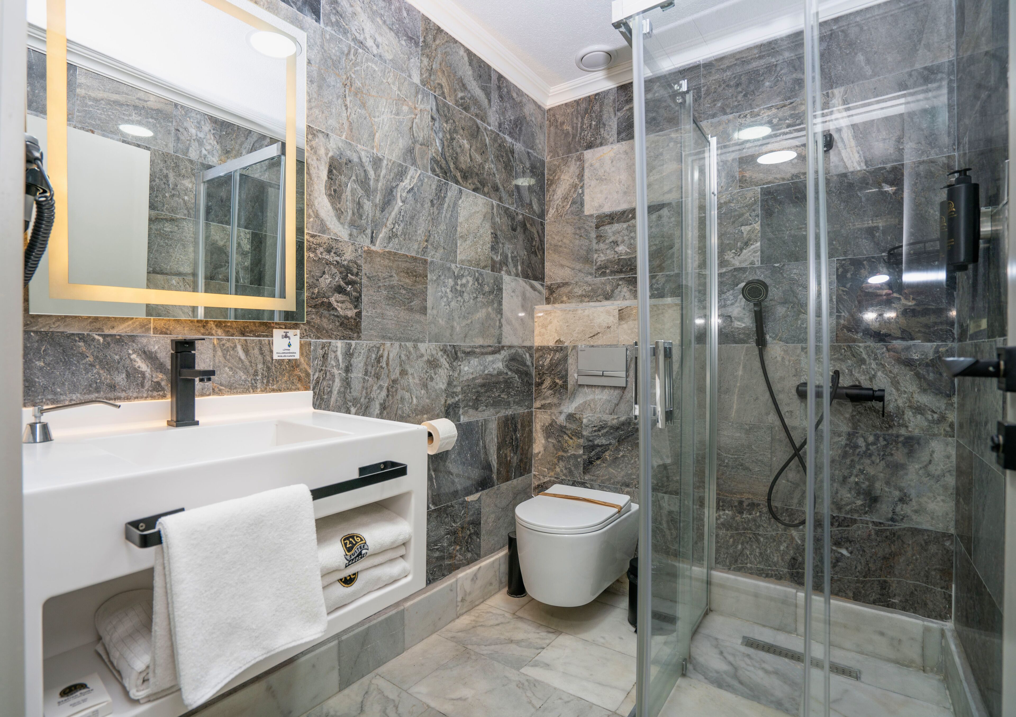 Un baño moderno con ducha de vidrio, paredes de mármol, lavabo blanco y un inodoro.