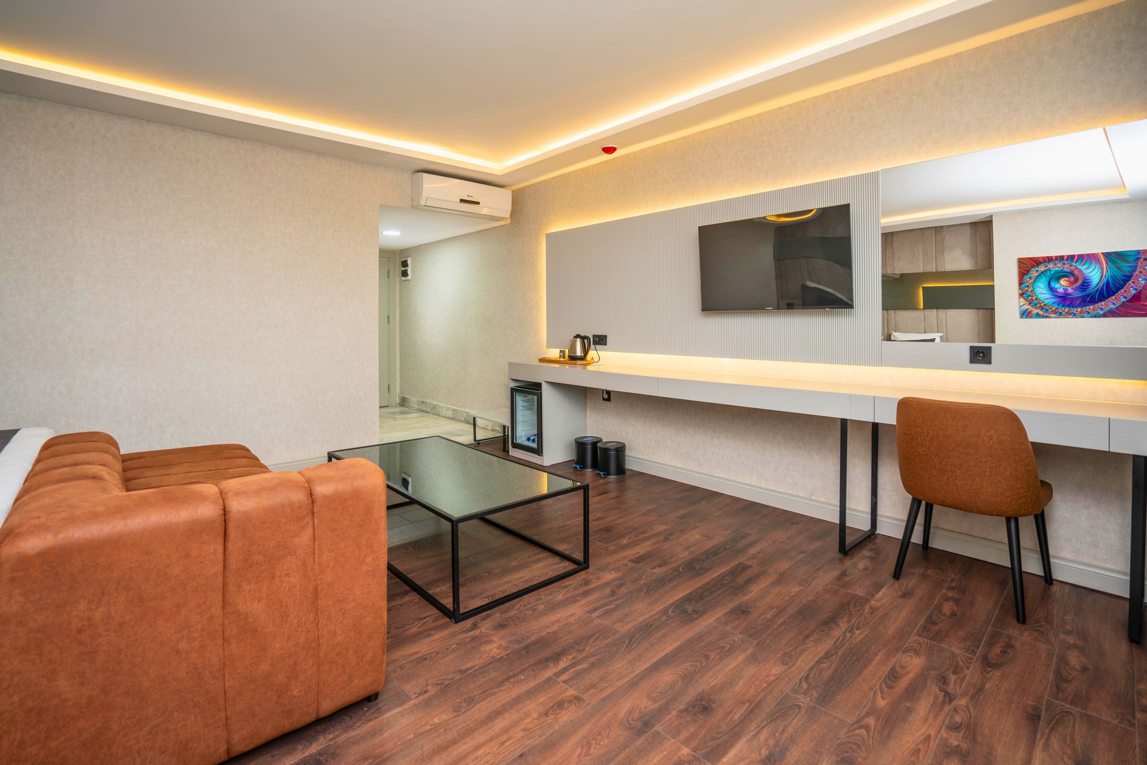 A modern hotel room with a wooden floor, a glass coffee table, a sofa, a desk with a chair, and a flat-screen TV mounted on the wall.