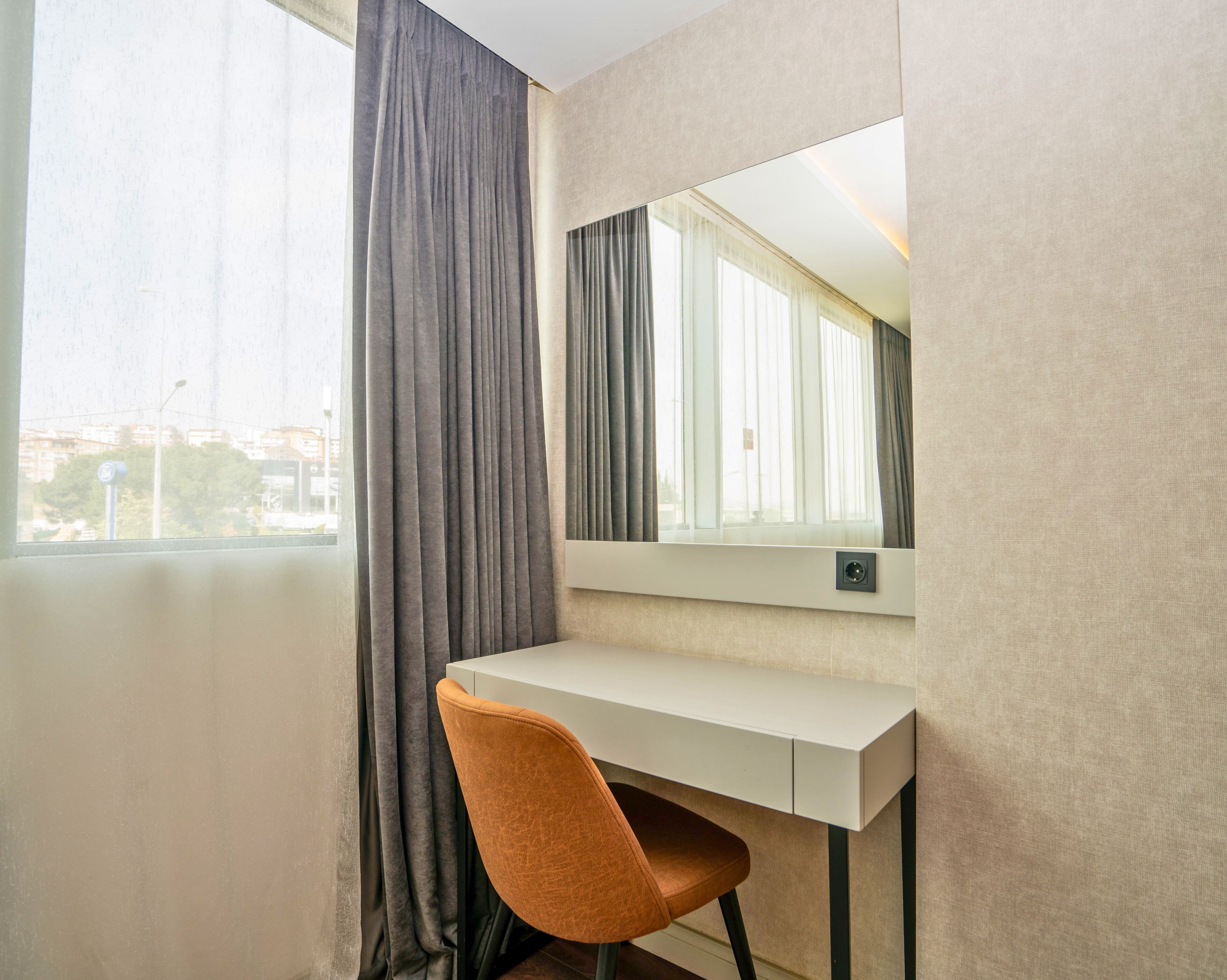 A hotel room with a white desk, a chair, a large mirror, and a window with curtains.