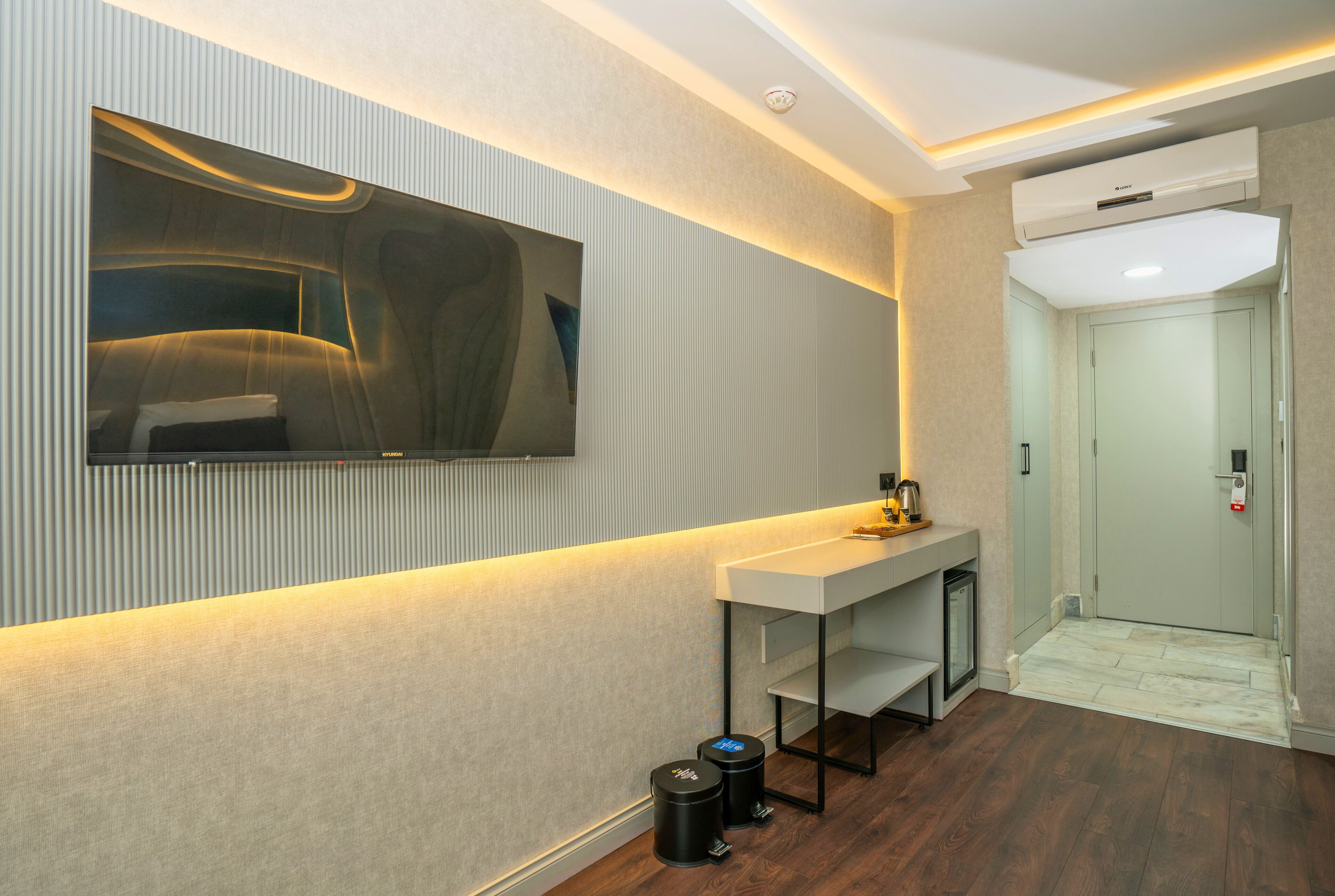 A modern hotel room with a large flat-screen TV mounted on the wall, a white console table, a small trash bin, and a door leading to the elevator.