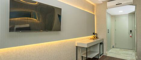 A modern hotel room with a large flat-screen TV mounted on the wall, a white console table, a small trash bin, and a door leading to the elevator.