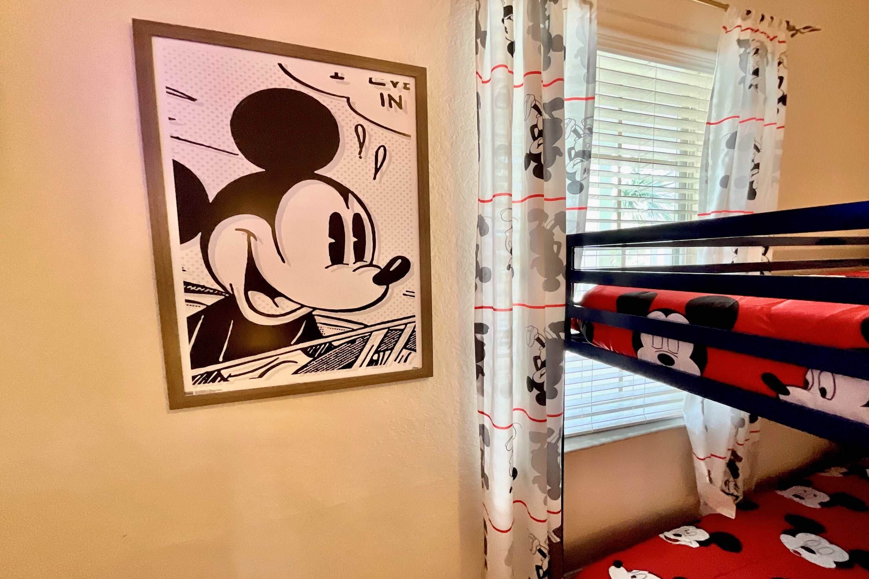 A framed cartoon character artwork on a wall, a window with curtains, and a bunk bed with Mickey Mouse-themed bedding.