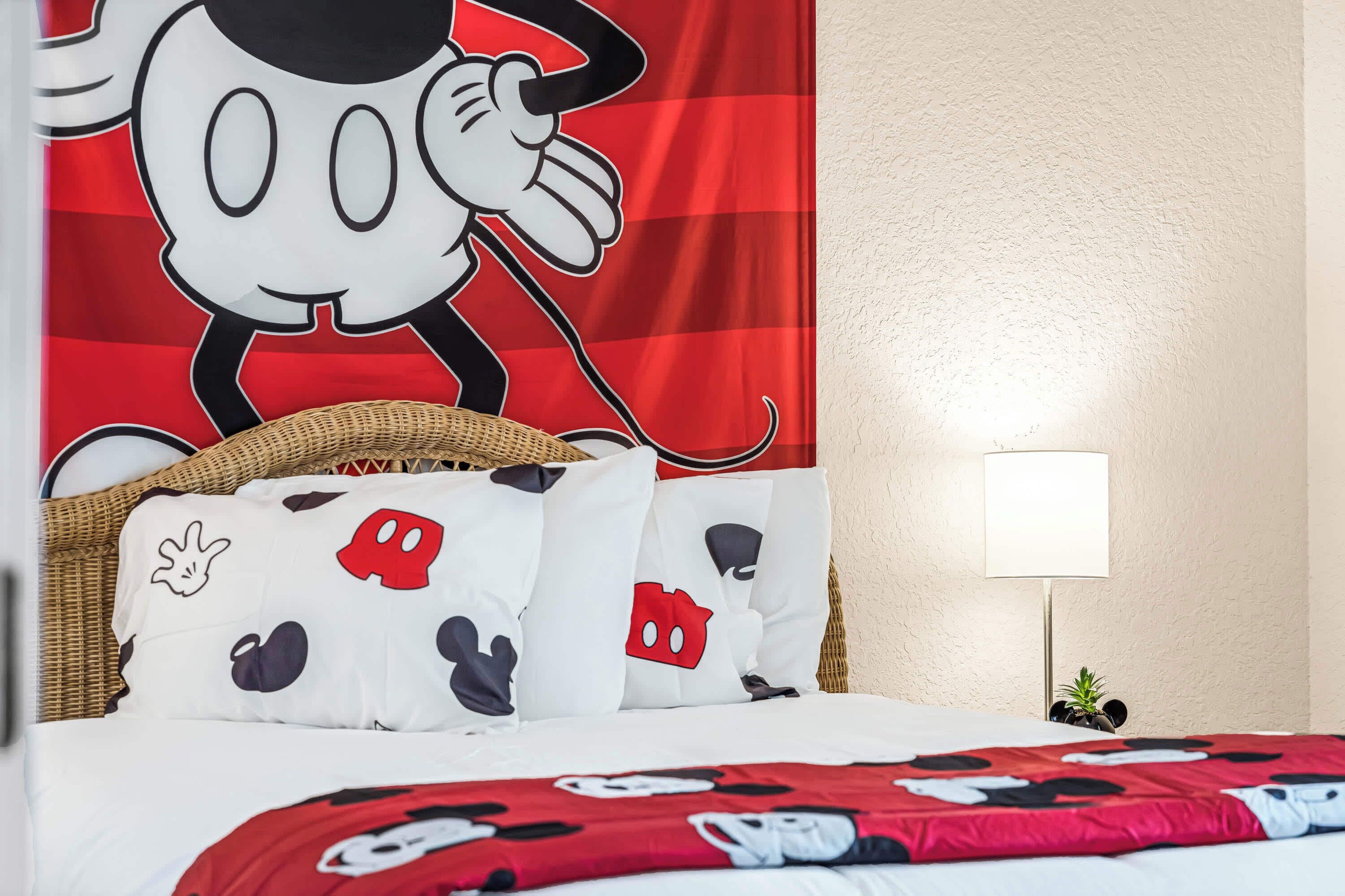 A hotel room with a large Mickey Mouse poster, a wicker headboard, a lamp, and a bed with Mickey Mouse themed bedding.