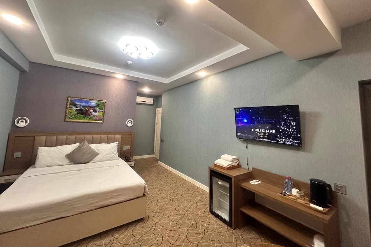 A hotel room with a bed, a television, a small desk with a coffee maker, and a wall-mounted TV displaying a cityscape.