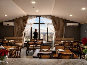 A restaurant with a view of the city, featuring tables and chairs, and a person standing by the window.