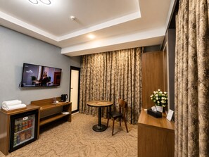 A hotel room with a television, a small refrigerator, a wooden bench, a round table with a chair, and a decorative plant. - Royal Meridien Hotel (Baku)