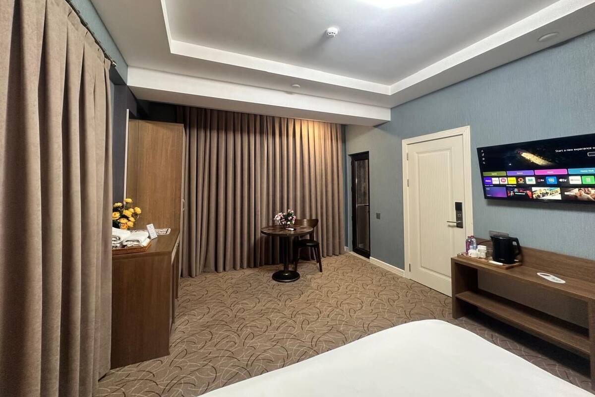 A hotel room with a large bed, a desk with a lamp, a chair, a TV mounted on the wall, and a curtain-covered window.