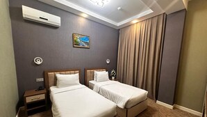 A hotel room with two beds, a nightstand, an air conditioning unit, a painting, and a window with curtains.