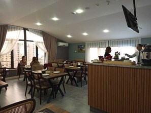 A dining area with multiple tables and chairs, a service counter, and large windows with curtains.