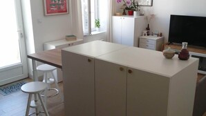 A modern kitchen with white cabinetry, a dining area with a table and two stools, and a large window with curtains. - Maison de Ville à Laleu - La Rochelle (La Rochelle)