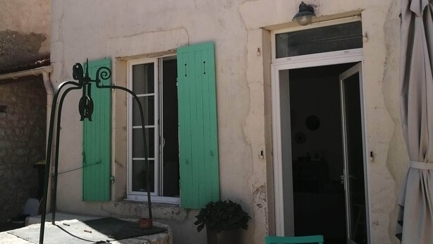 A building with multiple windows, some with green shutters, a small outdoor seating area with a table and chairs, and a potted plant. - Maison de Ville à Laleu - La Rochelle (La Rochelle)