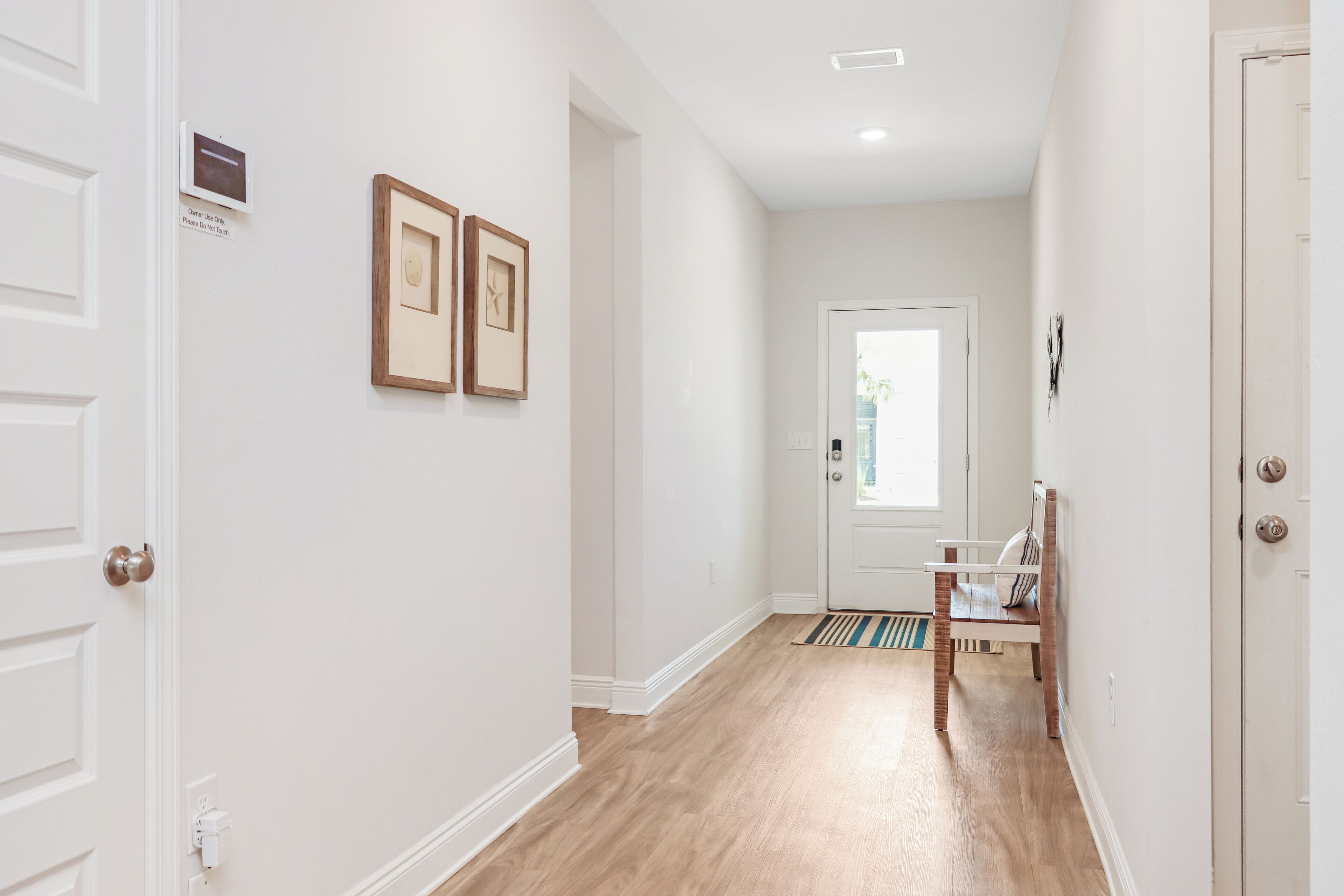 A hallway with a wooden floor, white walls, and a door at the end. There are two framed pictures on the wall and a small sign above the door.