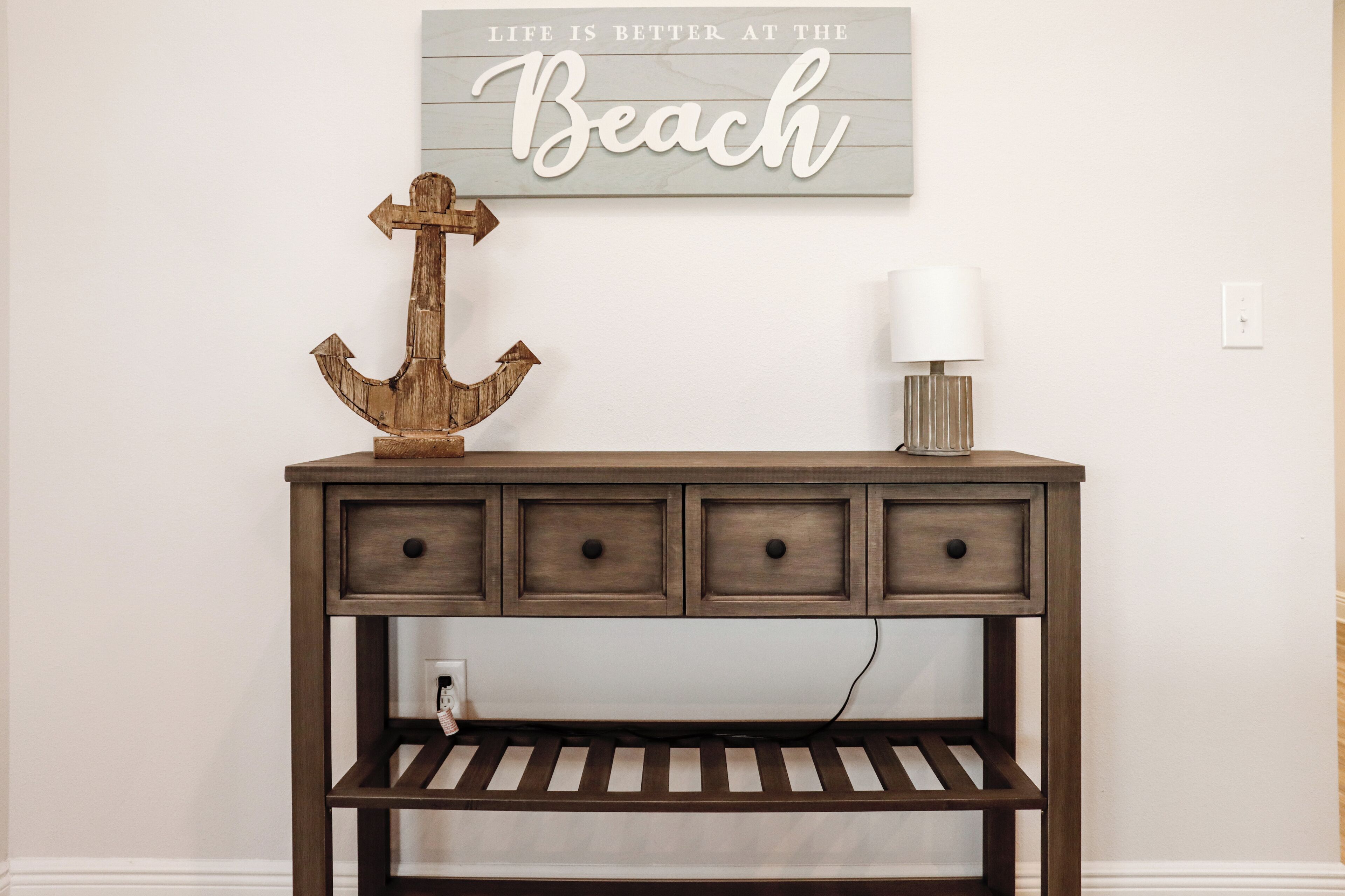 A wooden console table with drawers and a shelf, a decorative anchor, a wall sign saying "Life is better at the Beach", and a lamp.
