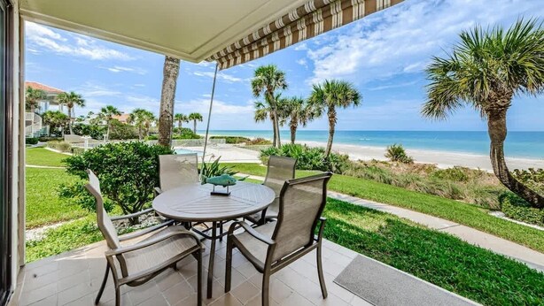 A covered patio with a table and chairs, overlooking a beach with palm trees and a clear sky.