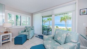 A living room with a sofa, ottoman, and a table with a lamp. There is a view of the beach through large windows.