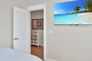Une chambre à coucher avec un lit, une porte blanche, une petite armoire et un téléviseur fixé au mur qui diffuse une scène de plage.
