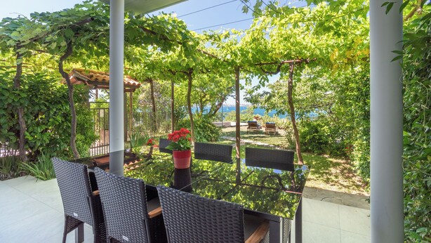 Eine Terrasse im Freien mit Korbmöbeln, einem mit Wein bewachsenen Pergola und Blick auf das Meer.