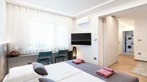 Ein modernes Hotelzimmer mit einem Bett, einem Schreibtisch mit Stuhl, einem an der Wand befestigten Fernseher und einer Waschmaschine im Hintergrund.