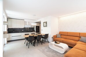 Eine moderne Küche mit weißen Schränken und einer schwarzen Arbeitsplatte, ein Essbereich mit einem Holztisch und schwarzen Stühlen sowie eine Wohnfläche mit einem orangefarbenen Sofa und einem weißen Couchtisch.