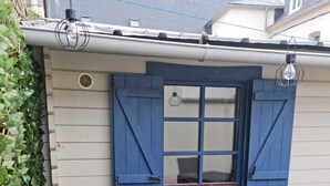 Un piccolo spazio all'aperto con un muro bianco, una finestra con persiane blu, una griglia da barbecue nera e un prato artificiale verde.
