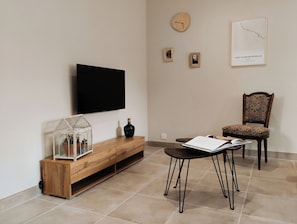 A living room with a TV mounted on the wall, a wooden console table with a glass terrarium, a chair with patterned upholstery, and a framed map on the wall.