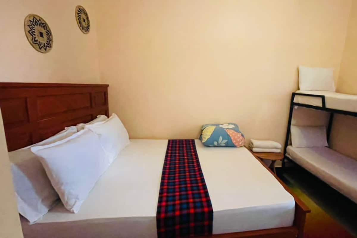 Un letto singolo con un poggiatesta in legno, una coperta a scacchi rossi e neri, e un cuscino. C'è un tavolino con un cuscino bianco e un cuscino azzurro con un motivo floreale. La stanza ha pareti spoglie e un pavimento chiaro.