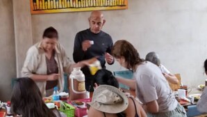 A group of people gathered around a table with food and drinks, in a room with a wooden ceiling and a colorful mural on the wall. - Maasai Hostel Tanzania (Arusha)